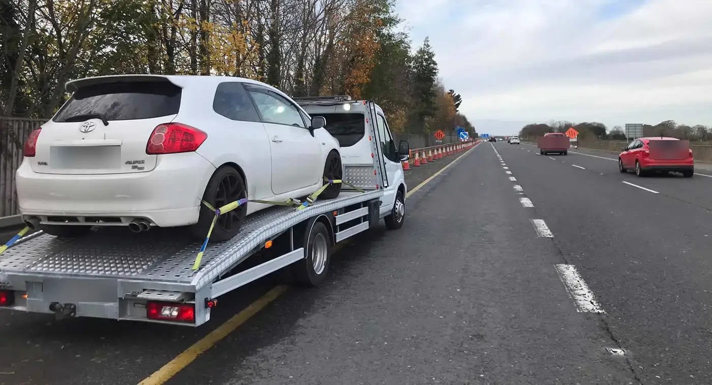 A small hatchback strapped onto a CC Recovery flatbed beside a busy dual carriageway