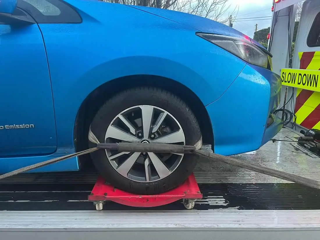 The front wheel of a blue electric car sitting on a red dolly, strapped to the deck of a CC Recovery truck