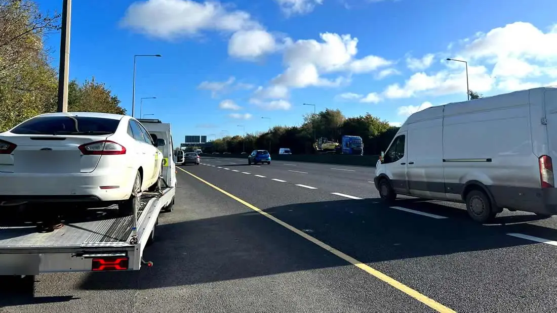 A CC Recovery truck carrying a white saloon on the hard shoulder of a dual carriageway under a blue sky