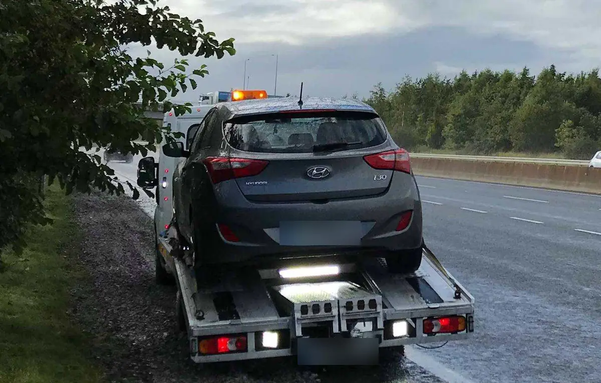 A grey hatchback loaded on a CC Recovery flatbed beside a dual carriageway, work lights on